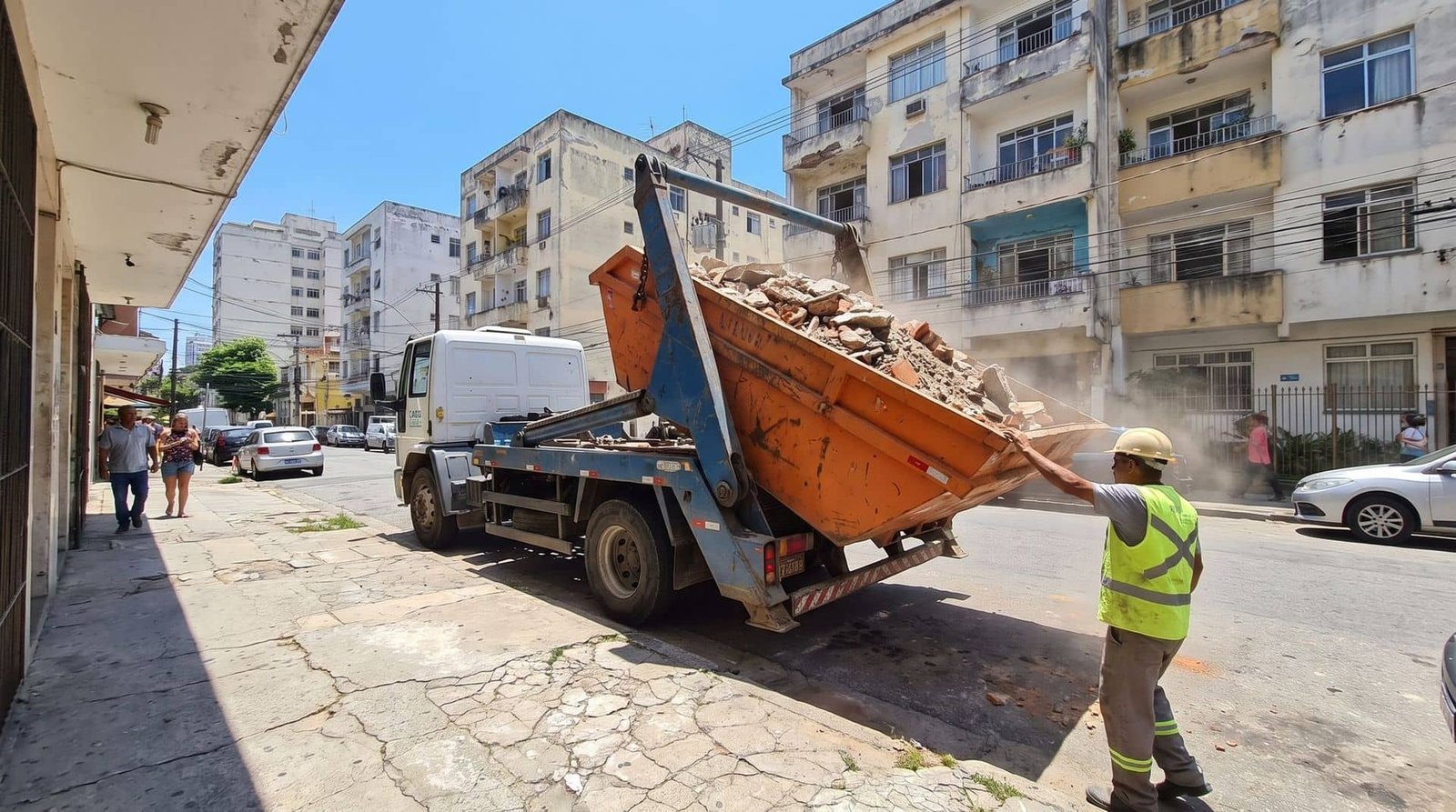 Caminhão poliguindaste içando uma caçamba cheia de entulho na rua
